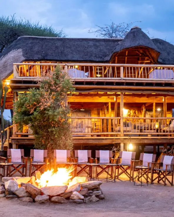 A campfire surrounded by chairs in front of Oliver's Camp glowing with warm lights at dusk