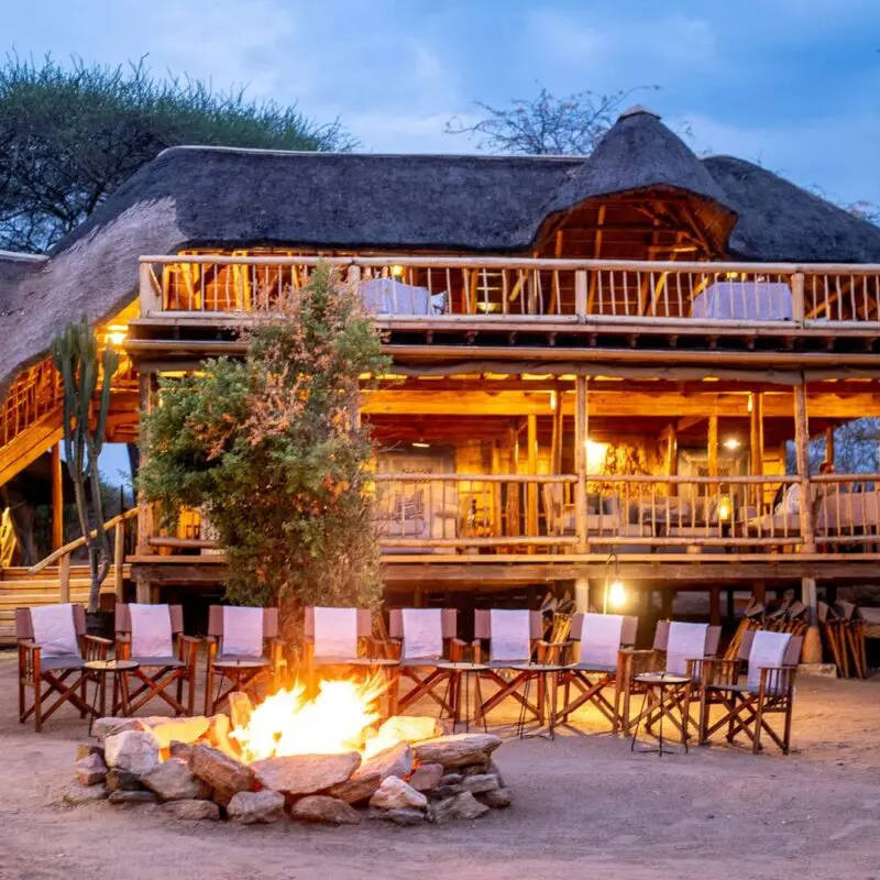 A campfire surrounded by chairs in front of Oliver's Camp glowing with warm lights at dusk