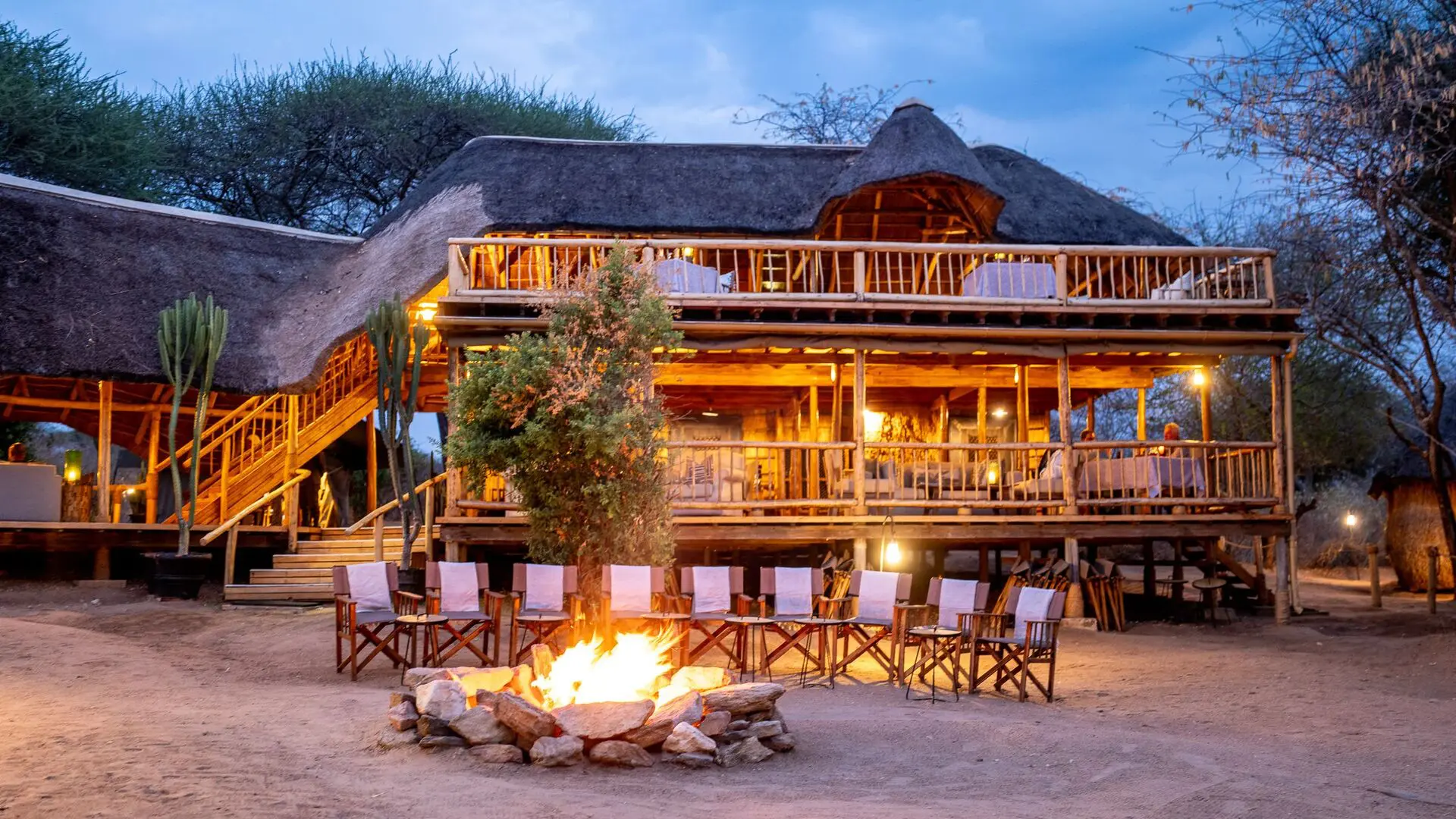 A campfire surrounded by chairs in front of Oliver's Camp glowing with warm lights at dusk