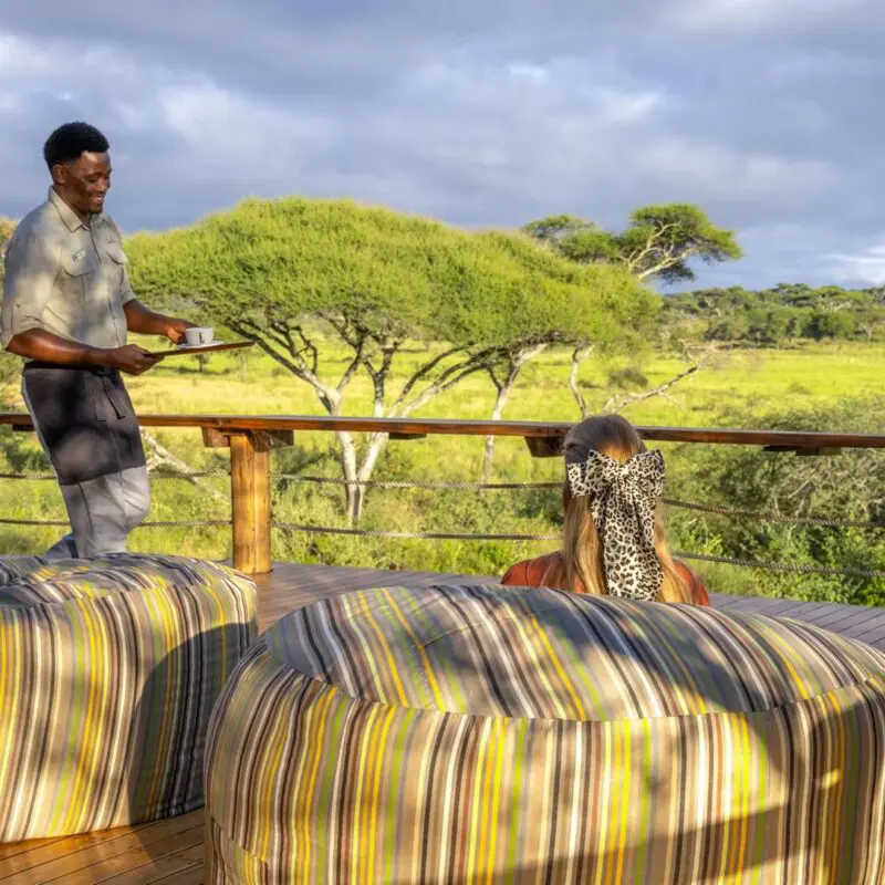 An Asilia staff member serves a drink to a female guest relaxing on a wooden deck, with comfortable seating and sweeping views of Tarangire National Park.