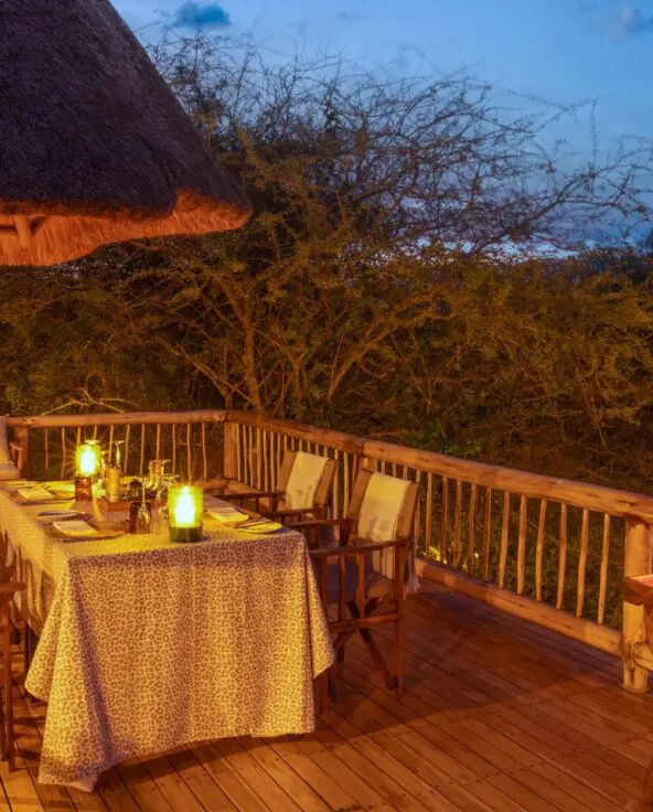 A wooden deck dining area with a table set for a meal, surrounded by lanterns and overlooking the bush at dusk