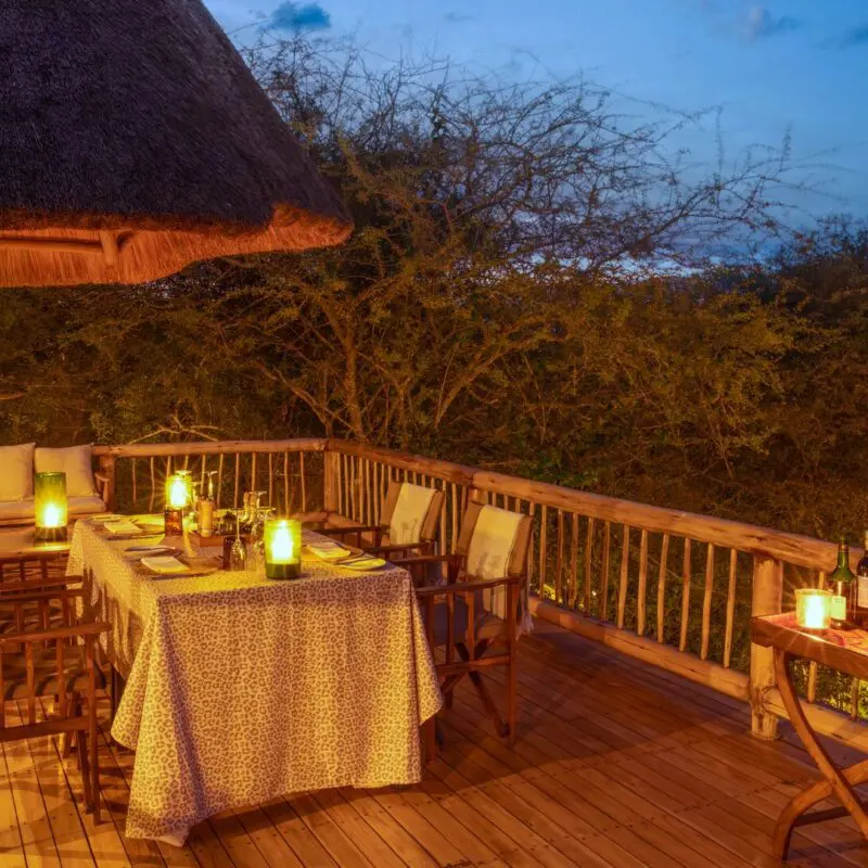 A wooden deck dining area with a table set for a meal, surrounded by lanterns and overlooking the bush at dusk