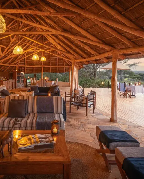 The spacious open-sided lounge and dining area at Oliver's Camp with warm lantern lighting, wooden beams, and expansive views across surrounding bushland at dusk.