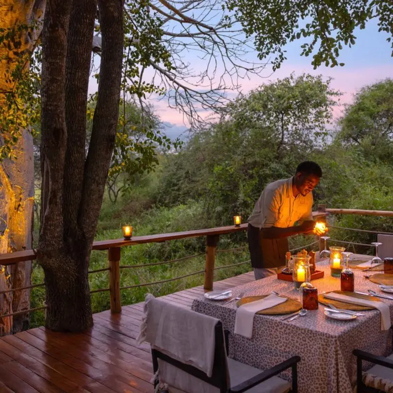 A staff member sets a candlelit dining table at Oliver's Camp on a wooden deck surrounded by trees, creating an intimate outdoor dinner setting at dusk.