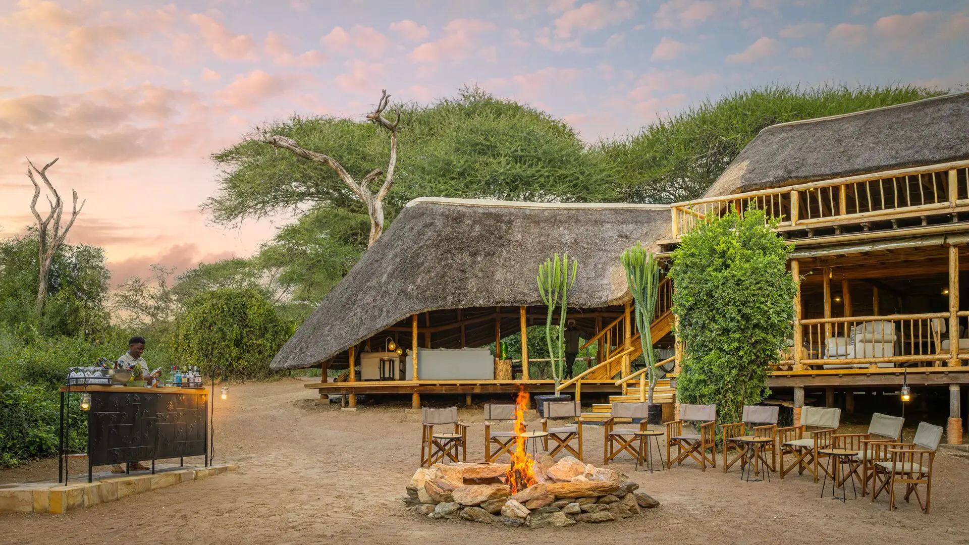 Outdoor area at Oliver's Camp with thatched buildings, a central fire pit surrounded by chairs, and a bar setup, set among trees at dusk with warm lantern light.