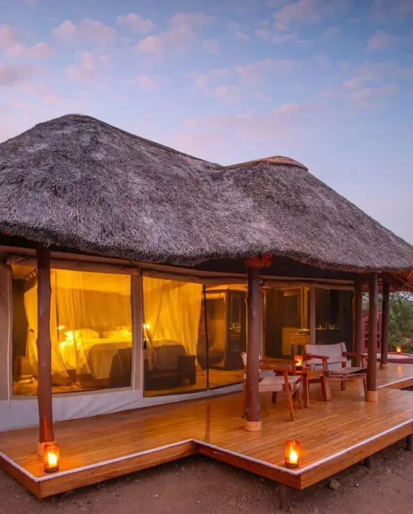The exterior of a luxury safari tent at Oliver's Camp with a thatched roof, wooden deck, and lanterns, softly lit at dusk and surrounded by natural bushland.