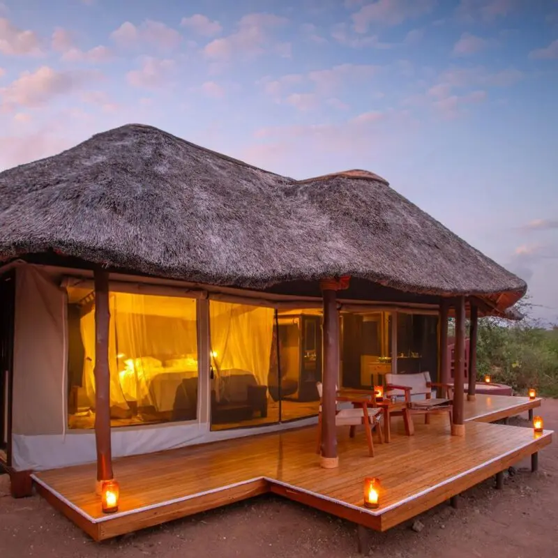 The exterior of a luxury safari tent at Oliver's Camp with a thatched roof, wooden deck, and lanterns, softly lit at dusk and surrounded by natural bushland.