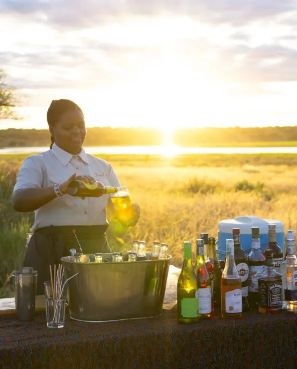 An Asilia staff member pours a drink at a bush bar setup with bottles and glassware during sunset