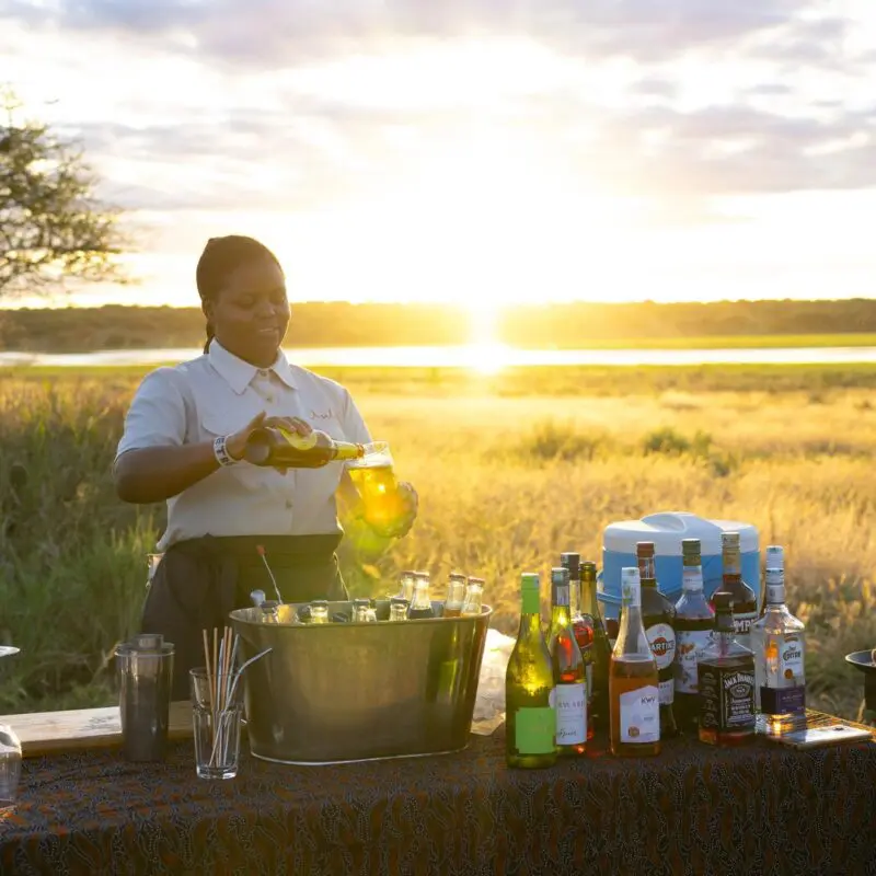 An Asilia staff member pours a drink at a bush bar setup with bottles and glassware during sunset