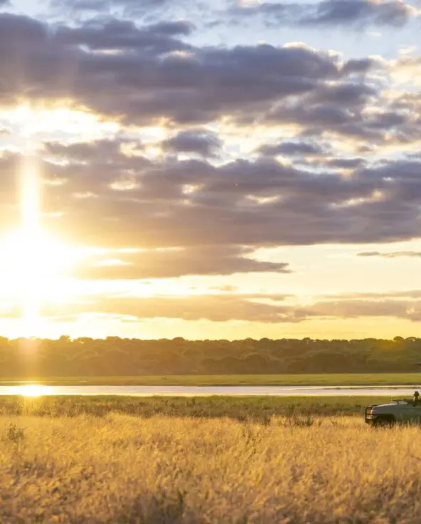 An Asilia safari vehicle pauses in golden grass at sunset, with guests watching the glowing sky reflected on a waterhole, framed by distant trees and layered clouds.