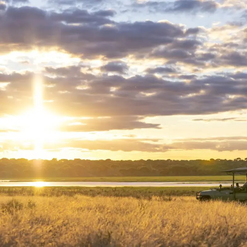 An Asilia safari vehicle pauses in golden grass at sunset, with guests watching the glowing sky reflected on a waterhole, framed by distant trees and layered clouds.