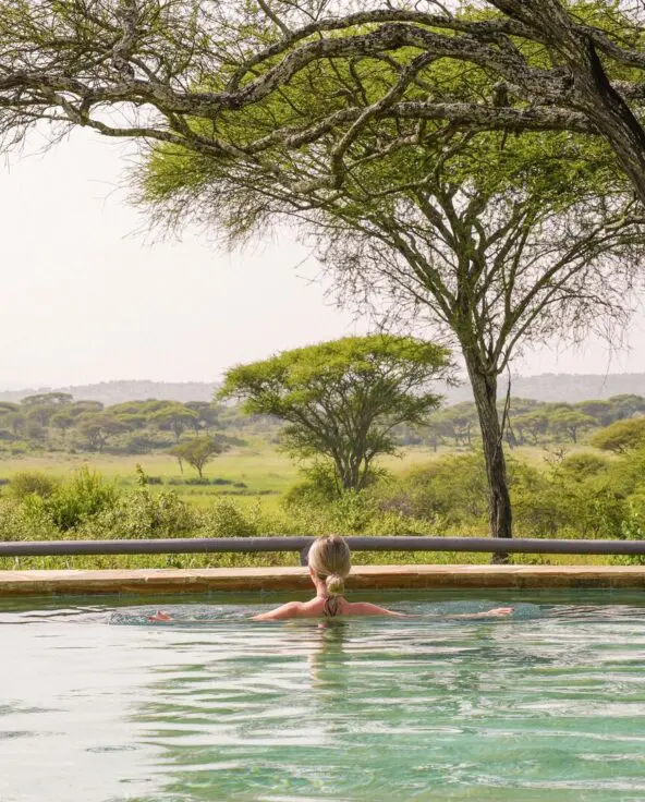 A guest relaxing in an infinity pool facing a wide savannah landscape with scattered acacia trees