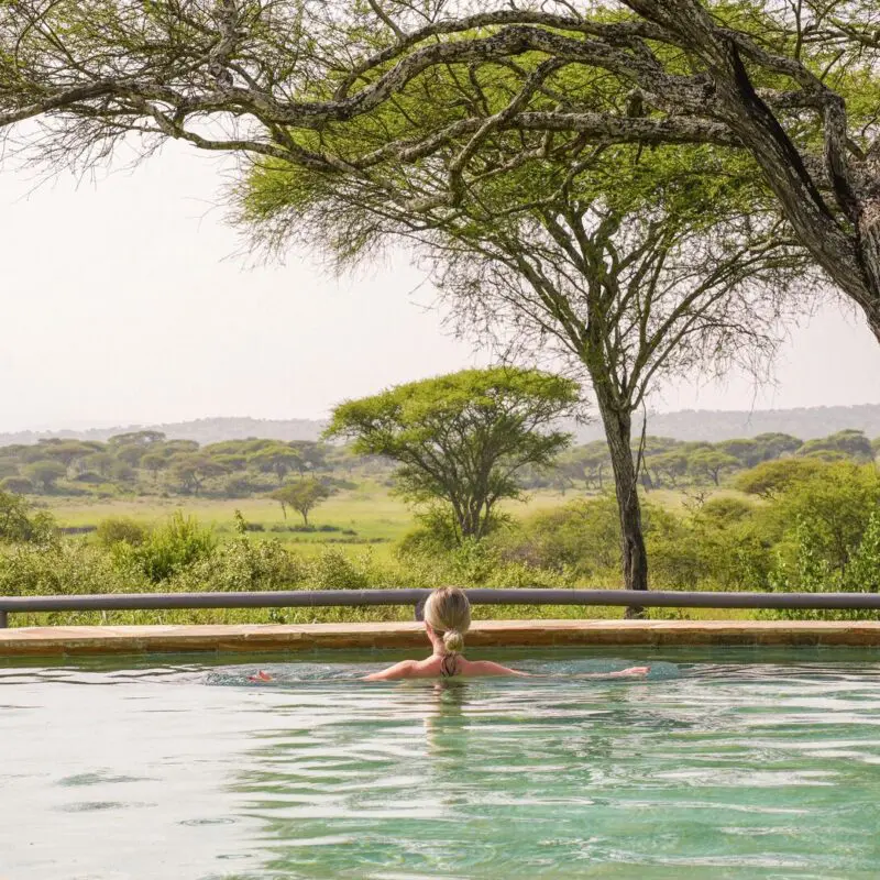 A guest relaxing in an infinity pool facing a wide savannah landscape with scattered acacia trees