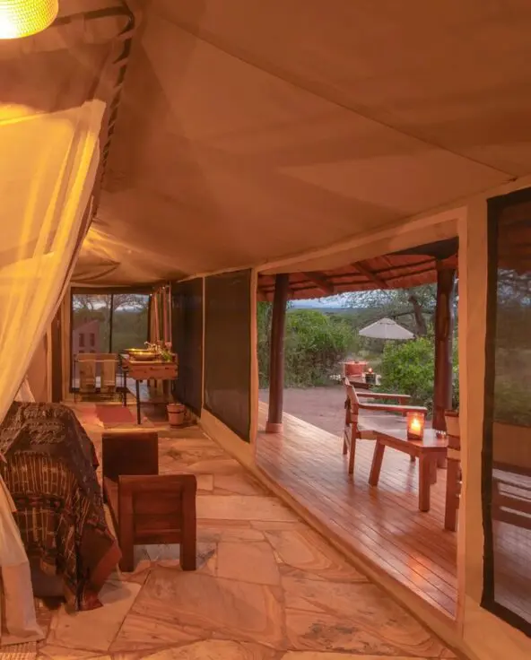 The interior of a luxury safari tent at Oliver's Camp with a draped mosquito-net bed, warm lighting, and stone floors, opening onto a private wooden deck overlooking bushland at dusk.