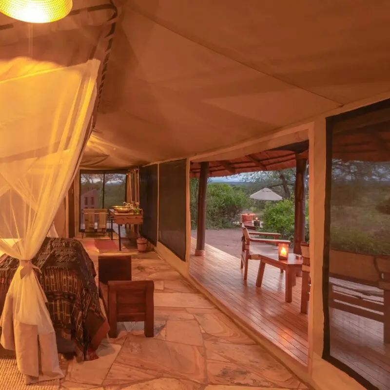 The interior of a luxury safari tent at Oliver's Camp with a draped mosquito-net bed, warm lighting, and stone floors, opening onto a private wooden deck overlooking bushland at dusk.