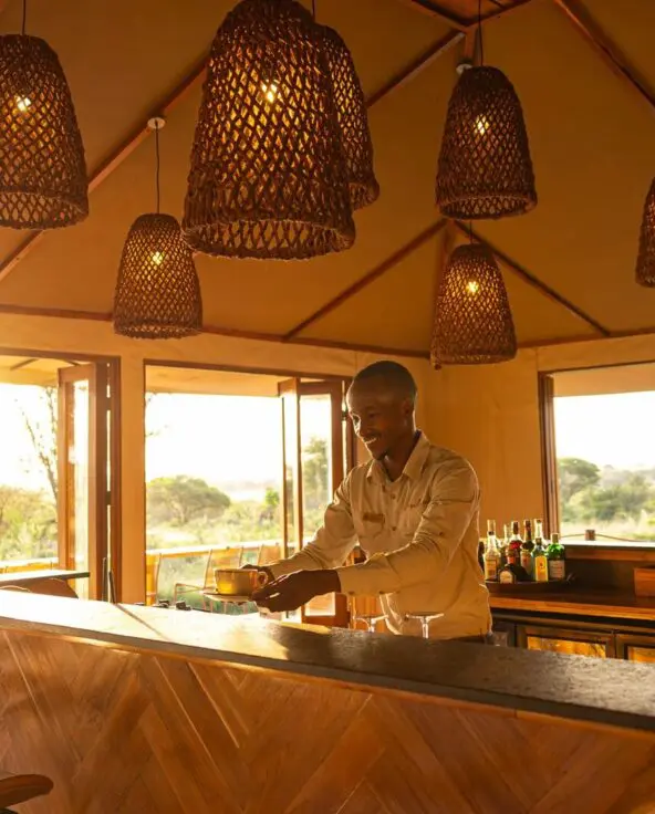 A smiling staff member prepares a drink behind a wooden safari lodge bar, warmly lit by woven pendant lights, with open windows revealing sunlit savannah and trees outside.