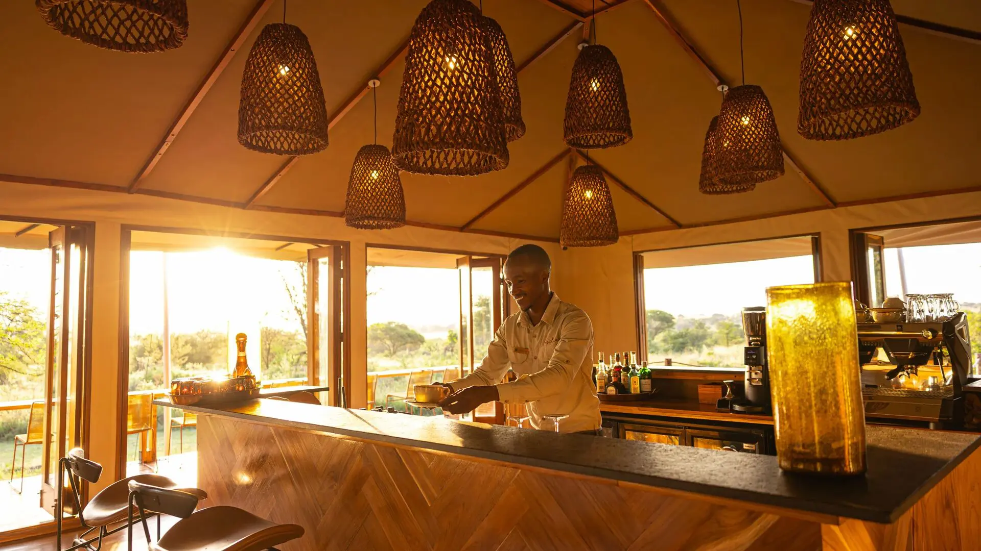 A smiling staff member prepares a drink behind a wooden safari lodge bar, warmly lit by woven pendant lights, with open windows revealing sunlit savannah and trees outside.