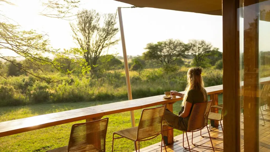 A guest sits at a wooden deck counter enjoying a cup of coffee, looking out over sunlit green savannah with scattered trees, as warm early-morning light filters through the camp veranda.