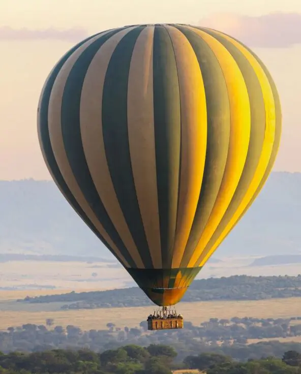 A large striped hot air balloon floats above the Serengeti plains at sunrise, carrying guests in a basket as golden light illuminates rolling grasslands, scattered trees, and distant hills.