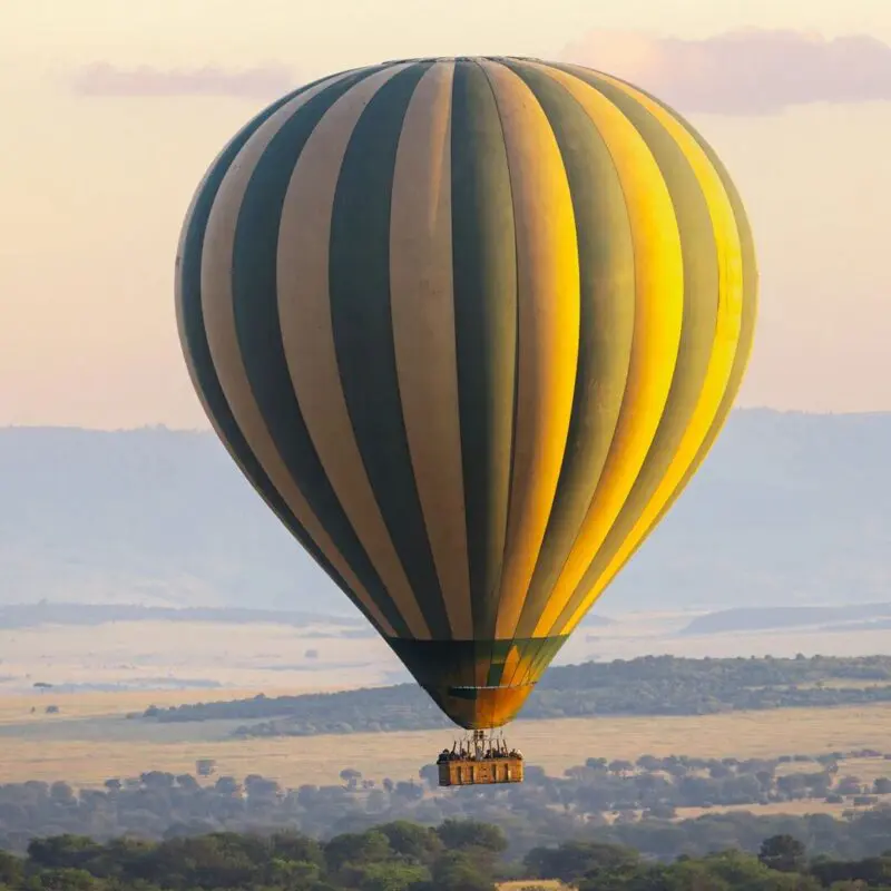 A large striped hot air balloon floats above the Serengeti plains at sunrise, carrying guests in a basket as golden light illuminates rolling grasslands, scattered trees, and distant hills.