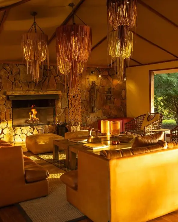 Sayari Camp's cosy lounge with leather sofas, a stone fireplace glowing with firelight, stacked firewood, woven pendant lights, and large open doors looking out onto green woodland.