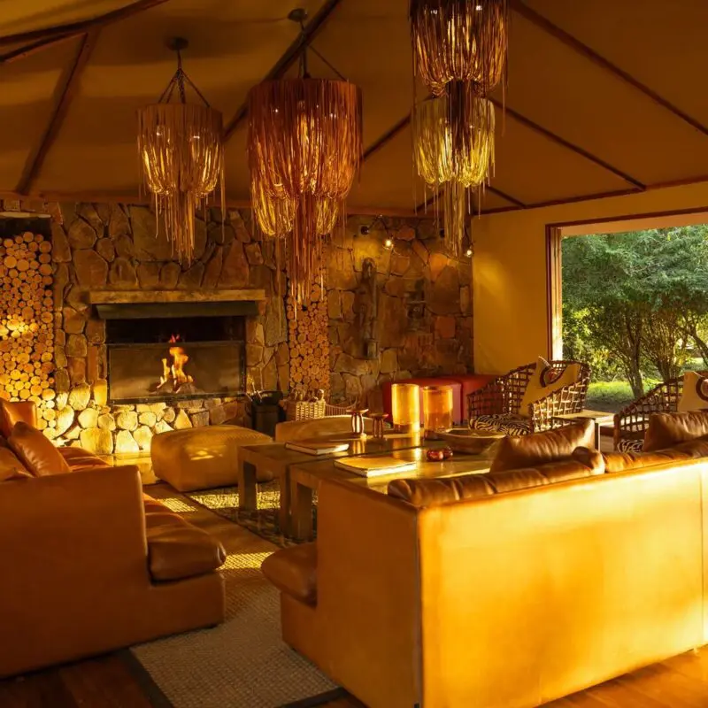Sayari Camp's cosy lounge with leather sofas, a stone fireplace glowing with firelight, stacked firewood, woven pendant lights, and large open doors looking out onto green woodland.