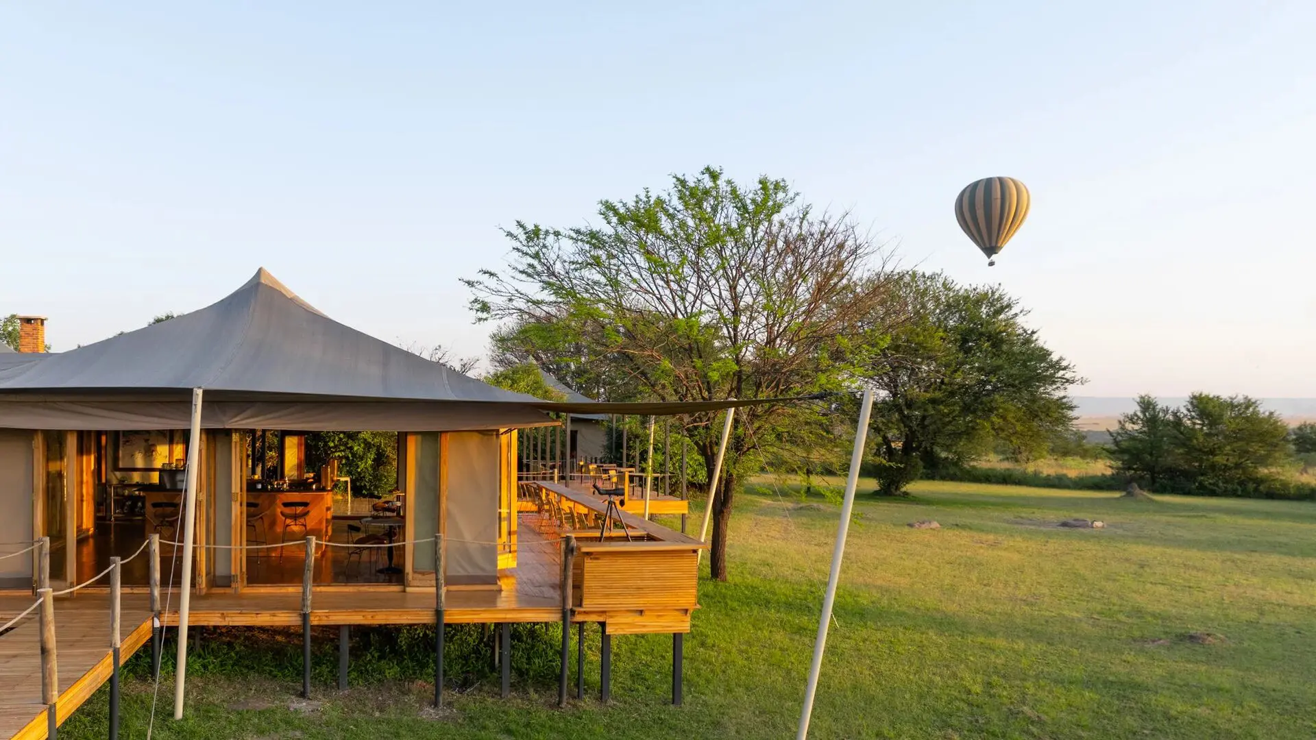 Sayari Camp’s raised lounge and deck overlooking open Serengeti grassland, with a hot air balloon floating in the distance during early morning light, framed by trees and clear skies.