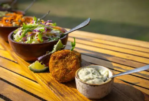 Close-up of a plated lunch at Sayari Camp on a wooden table, featuring golden croquettes with dipping sauce, fresh mixed salads in wooden bowls, and vibrant vegetables, set outdoors in warm natural light.