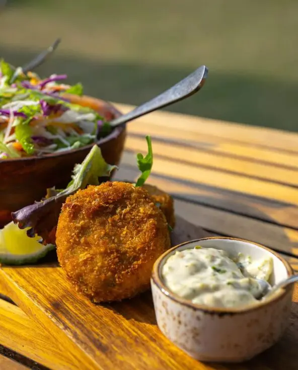 Close-up of a plated lunch at Sayari Camp on a wooden table, featuring golden croquettes with dipping sauce, fresh mixed salads in wooden bowls, and vibrant vegetables, set outdoors in warm natural light.