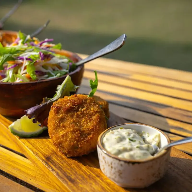 Close-up of a plated lunch at Sayari Camp on a wooden table, featuring golden croquettes with dipping sauce, fresh mixed salads in wooden bowls, and vibrant vegetables, set outdoors in warm natural light.
