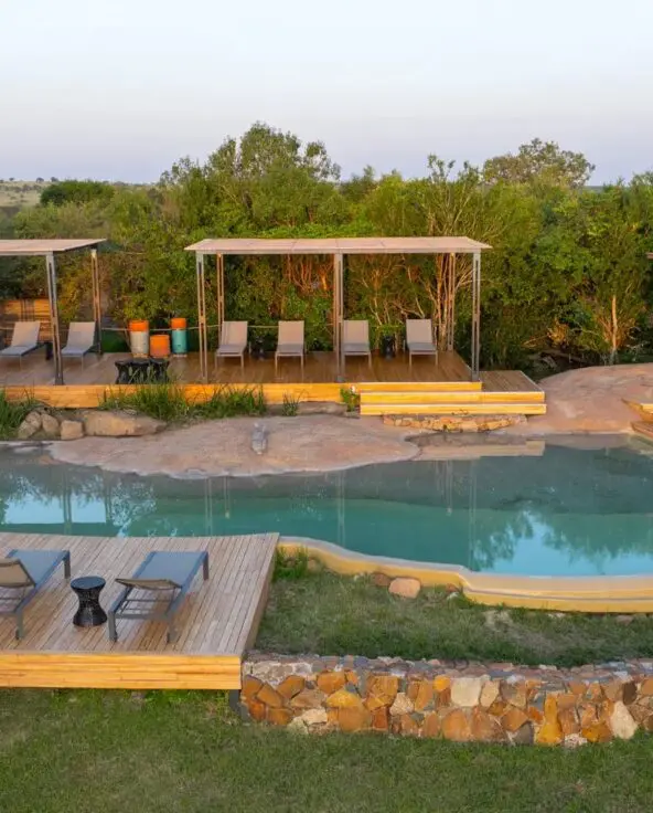 Sayari Camp’s natural stone and wood swimming pool surrounded by sun loungers and shaded decks, set among green trees and open Serengeti plains in soft morning light.