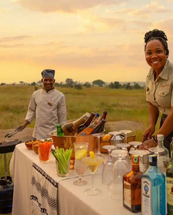 Two Sayari Camp staff prepare a classic safari sundowner setup on the open plains, with a table of drinks, glasses, and snacks at sunset, golden grasslands stretching into the distance beneath a warm evening sky.