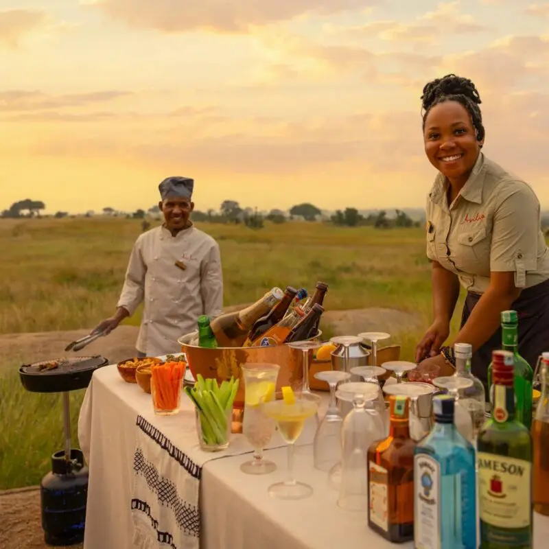 Two Sayari Camp staff prepare a classic safari sundowner setup on the open plains, with a table of drinks, glasses, and snacks at sunset, golden grasslands stretching into the distance beneath a warm evening sky.