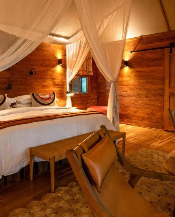 Interior of a Sayari Retreat double room featuring a king-size bed draped with mosquito netting, rich wooden walls and floors, soft ambient lighting, leather lounge chairs, a writing desk with round mirror, and a calm, intimate safari-lodge atmosphere.