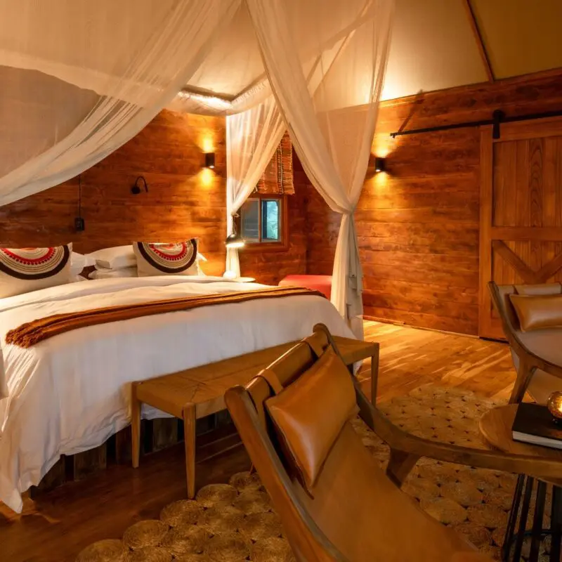 Interior of a Sayari Retreat double room featuring a king-size bed draped with mosquito netting, rich wooden walls and floors, soft ambient lighting, leather lounge chairs, a writing desk with round mirror, and a calm, intimate safari-lodge atmosphere.