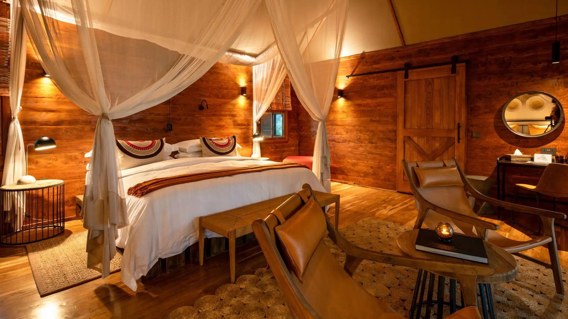 Interior of a Sayari Retreat double room featuring a king-size bed draped with mosquito netting, rich wooden walls and floors, soft ambient lighting, leather lounge chairs, a writing desk with round mirror, and a calm, intimate safari-lodge atmosphere.