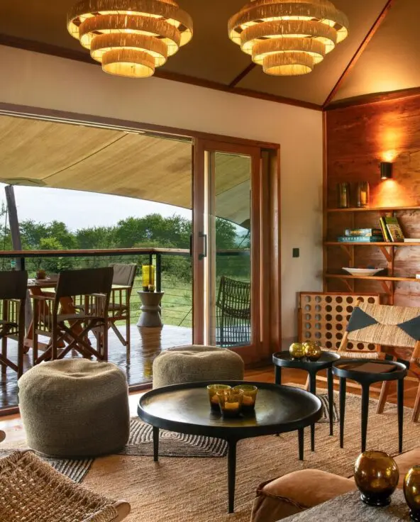 The warm, wood-paneled lounge at Sayari Retreat with plush seating, woven chairs, and soft lighting opens through glass doors onto a covered deck with a dining table, overlooking green savannah trees and open wilderness beyond.