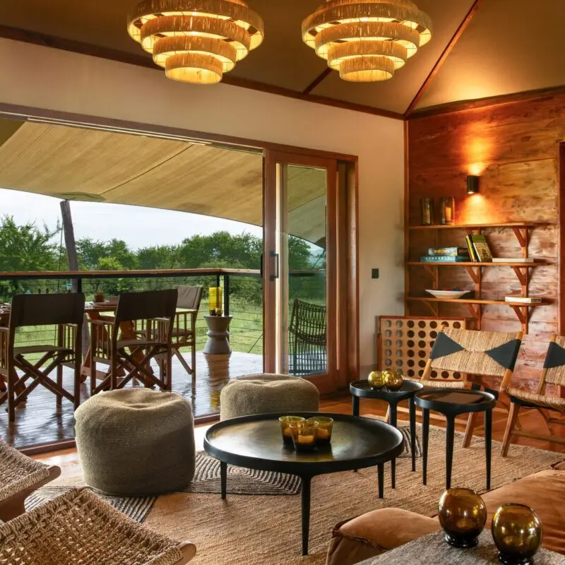The warm, wood-paneled lounge at Sayari Retreat with plush seating, woven chairs, and soft lighting opens through glass doors onto a covered deck with a dining table, overlooking green savannah trees and open wilderness beyond.