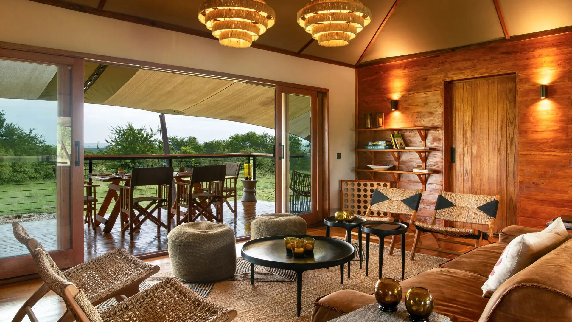 The warm, wood-paneled lounge at Sayari Retreat with plush seating, woven chairs, and soft lighting opens through glass doors onto a covered deck with a dining table, overlooking green savannah trees and open wilderness beyond.