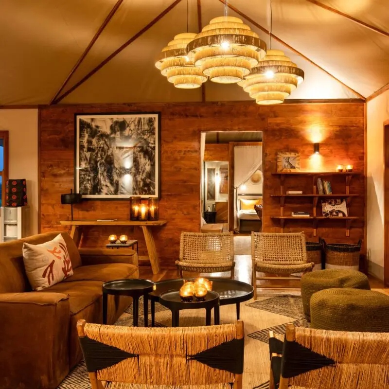 Interior of a cosy private lounge at Sayari Retreat, featuring warm wooden walls, woven chairs, a plush sofa, soft ambient lighting, and large glass doors opening onto a deck, creating an intimate safari-lodge atmosphere.