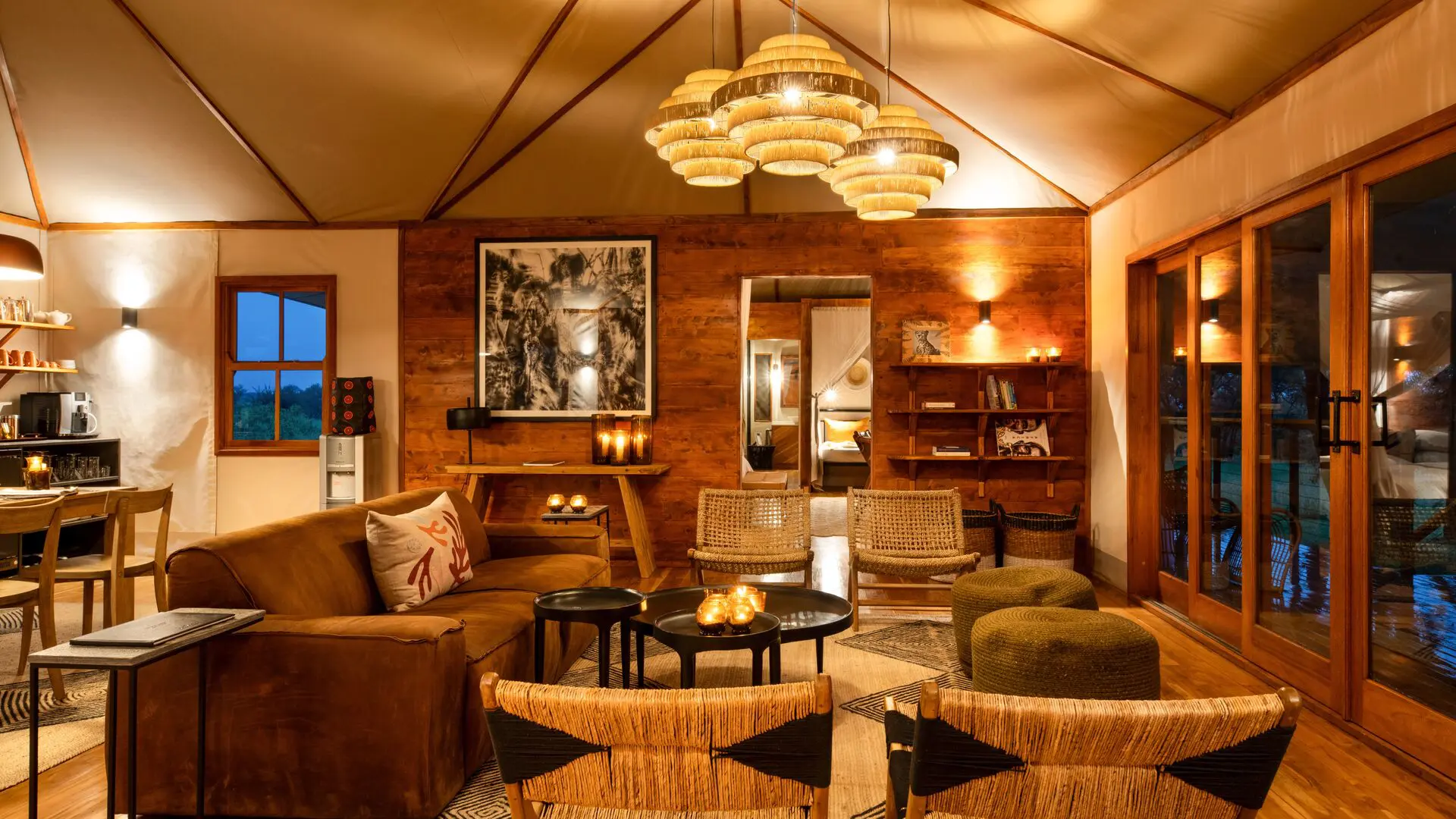 Interior of a cosy private lounge at Sayari Retreat, featuring warm wooden walls, woven chairs, a plush sofa, soft ambient lighting, and large glass doors opening onto a deck, creating an intimate safari-lodge atmosphere.
