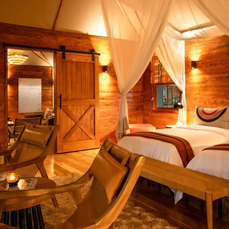 Interior of a twin bedroom at Sayari Retreat featuring two canopy beds with white linens, warm wooden walls, soft ambient lighting, leather lounge chairs, and a cosy, refined safari-lodge design.