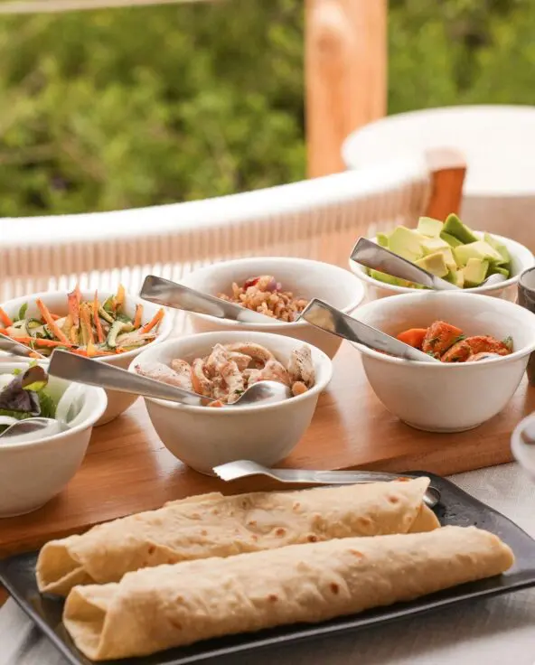 Outdoor lunch spread on a wooden table featuring wraps, bowls of salads, grains, chicken, avocado, sauces, and glasses of rosé wine, set on a deck overlooking lush green forest.