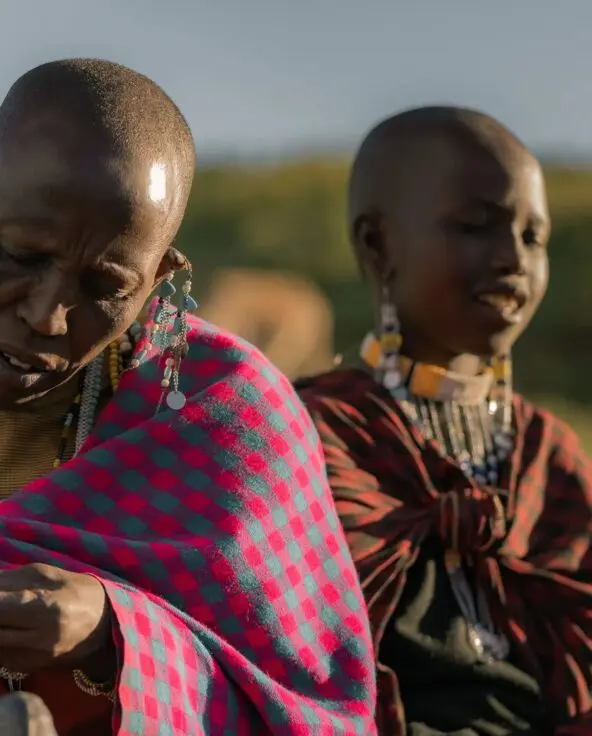 Two Maasai women seated outdoors on open grassland, wearing traditional red and patterned shukas and beaded jewelry, carefully handcrafting beadwork together in warm afternoon light.