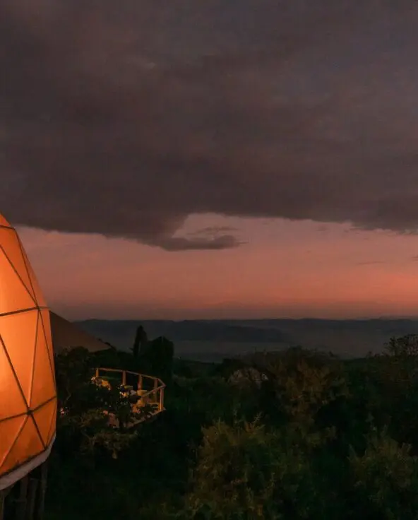 A softly lit geodesic dome at The Highlands Camp at dusk, glowing warmly against dark clouds and a fading pink horizon, overlooking forested slopes and distant highlands.