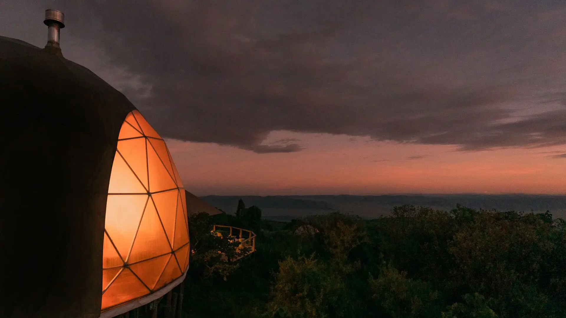 A softly lit geodesic dome at The Highlands Camp at dusk, glowing warmly against dark clouds and a fading pink horizon, overlooking forested slopes and distant highlands.