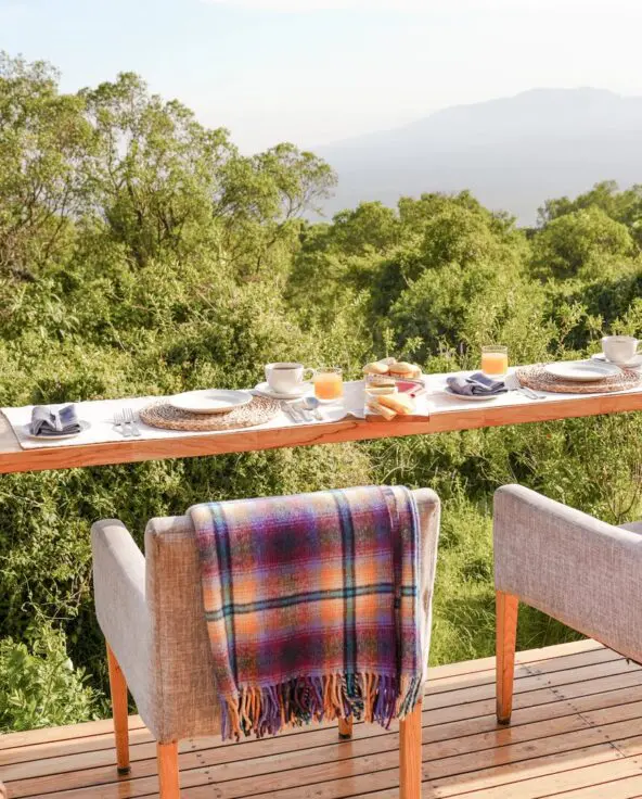 Outdoor dining setup at The Highlands on a wooden deck with a long counter table set for breakfast, cushioned chairs draped with blankets, and views over dense green forest toward distant mountains under soft morning light.