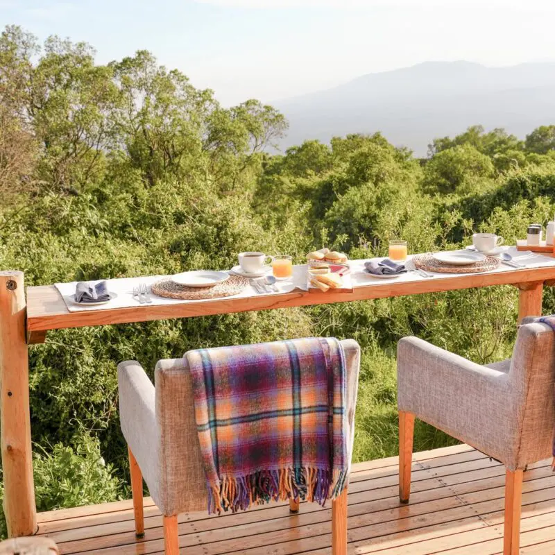 Outdoor dining setup at The Highlands on a wooden deck with a long counter table set for breakfast, cushioned chairs draped with blankets, and views over dense green forest toward distant mountains under soft morning light.