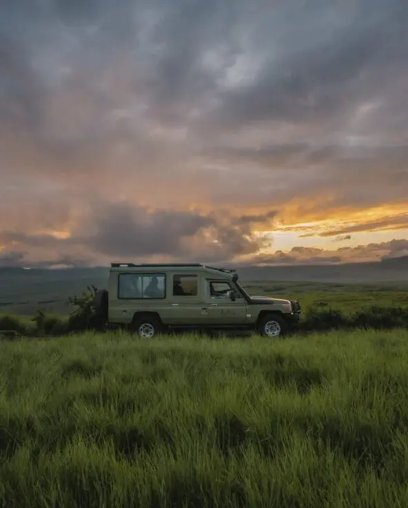 An Asilia safari vehicle parked on a grassy plain at sunset, with dramatic clouds glowing orange and purple over rolling highland hills and distant mountains.