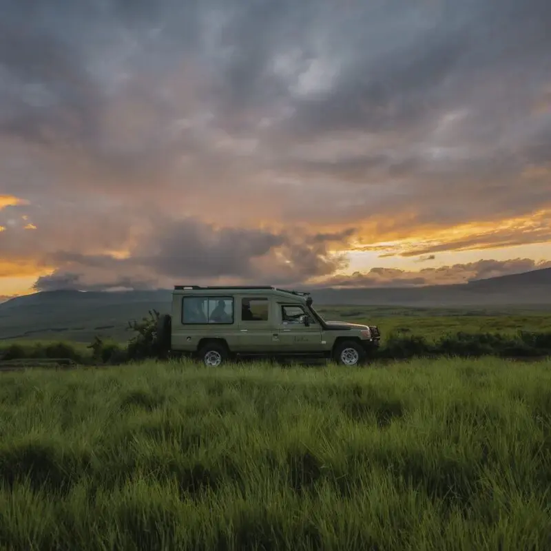 An Asilia safari vehicle parked on a grassy plain at sunset, with dramatic clouds glowing orange and purple over rolling highland hills and distant mountains.